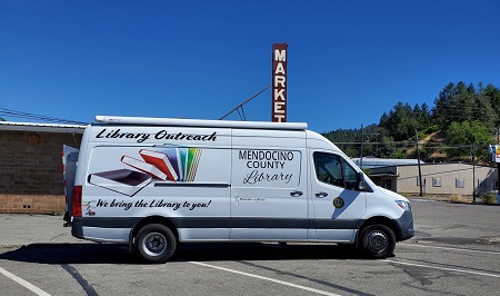 Outreach Van in a parking lot with a sign reading market in the background.